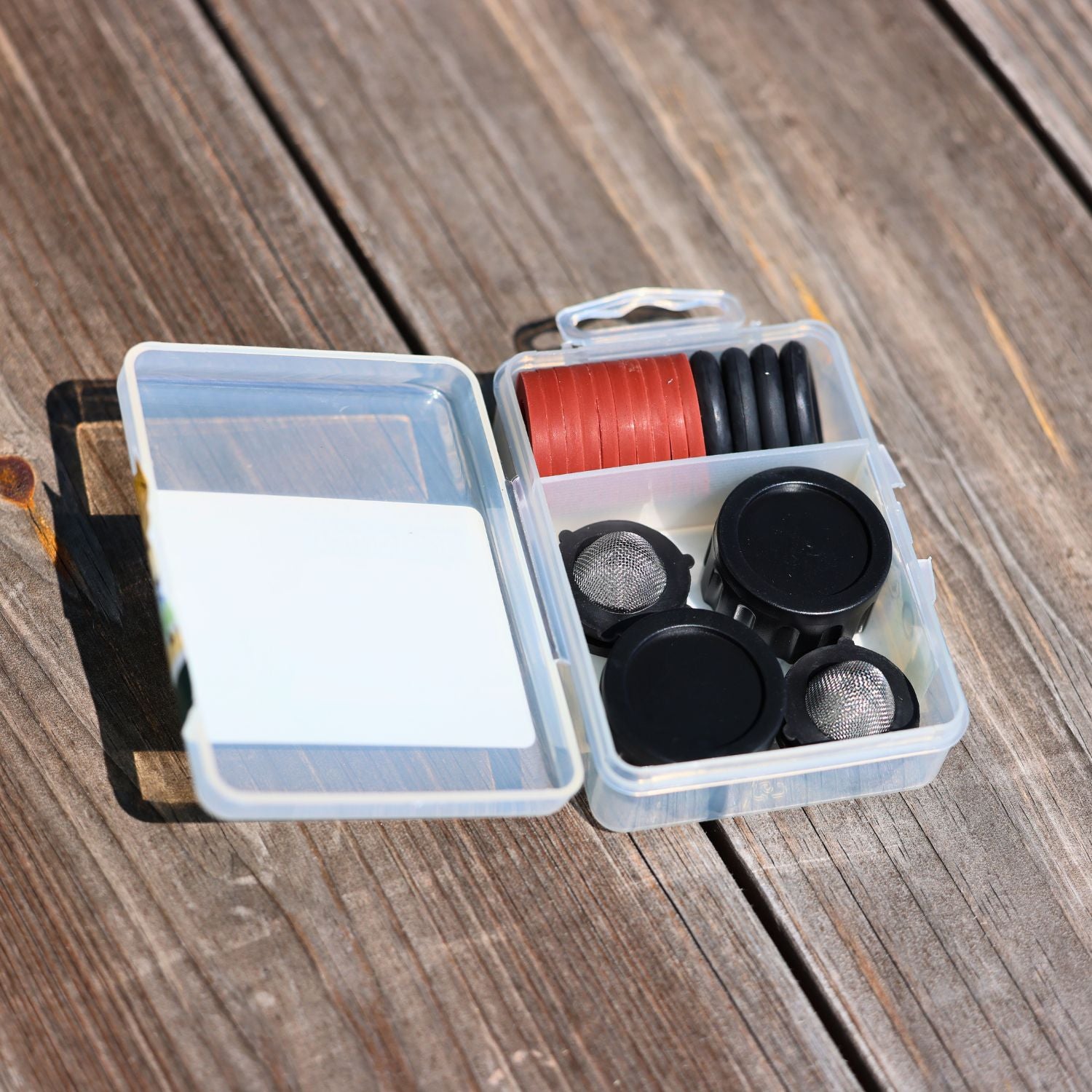 A clear plastic box with various camera lens filters and red thread rests on a wooden surface, open and organized, showcasing photography equipment.
