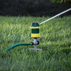 A sprinkler is attached to a green hose, spraying water onto lush grass. The background is slightly shaded, indicating it might be early morning or late afternoon.