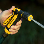 A person is holding a yellow and black garden hose nozzle emitting a strong, focused stream of water, set against a blurred green background.