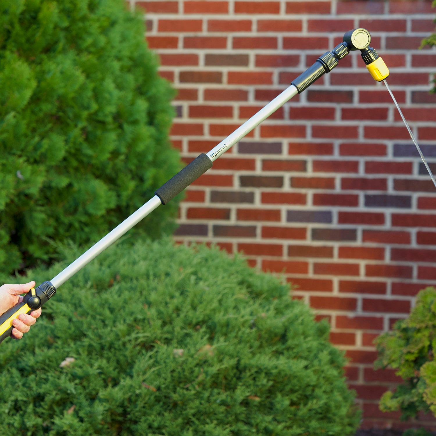 A person's hand is holding an extended water hose nozzle against a background of a brick wall and green shrubbery. The device is likely used for gardening.