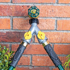 A Y split garden hose connector mounted on a red brick wall, featuring a gray body with two yellow valve controls with a hose connected to each valve.