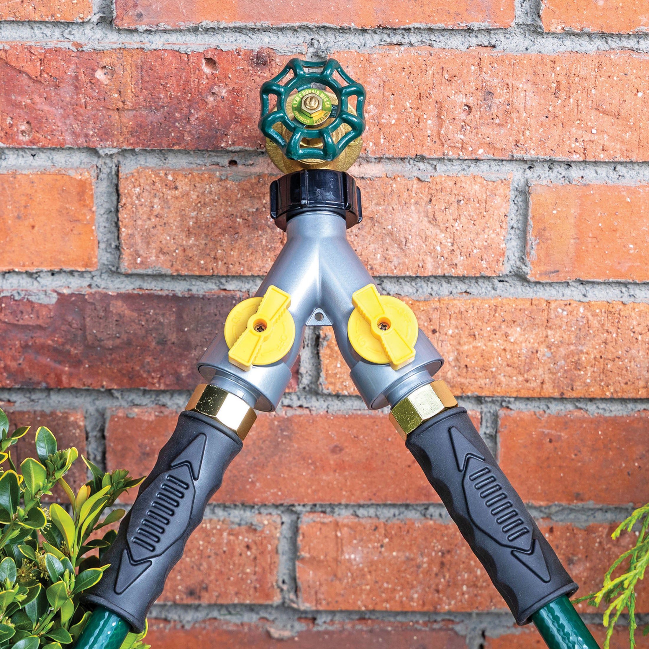 A Y split garden hose connector mounted on a red brick wall, featuring a gray body with two yellow valve controls with a hose connected to each valve.
