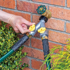 A Y split garden hose connector mounted on a red brick wall, featuring a gray body with a hand reaching out to turn one of two yellow valve controls .
