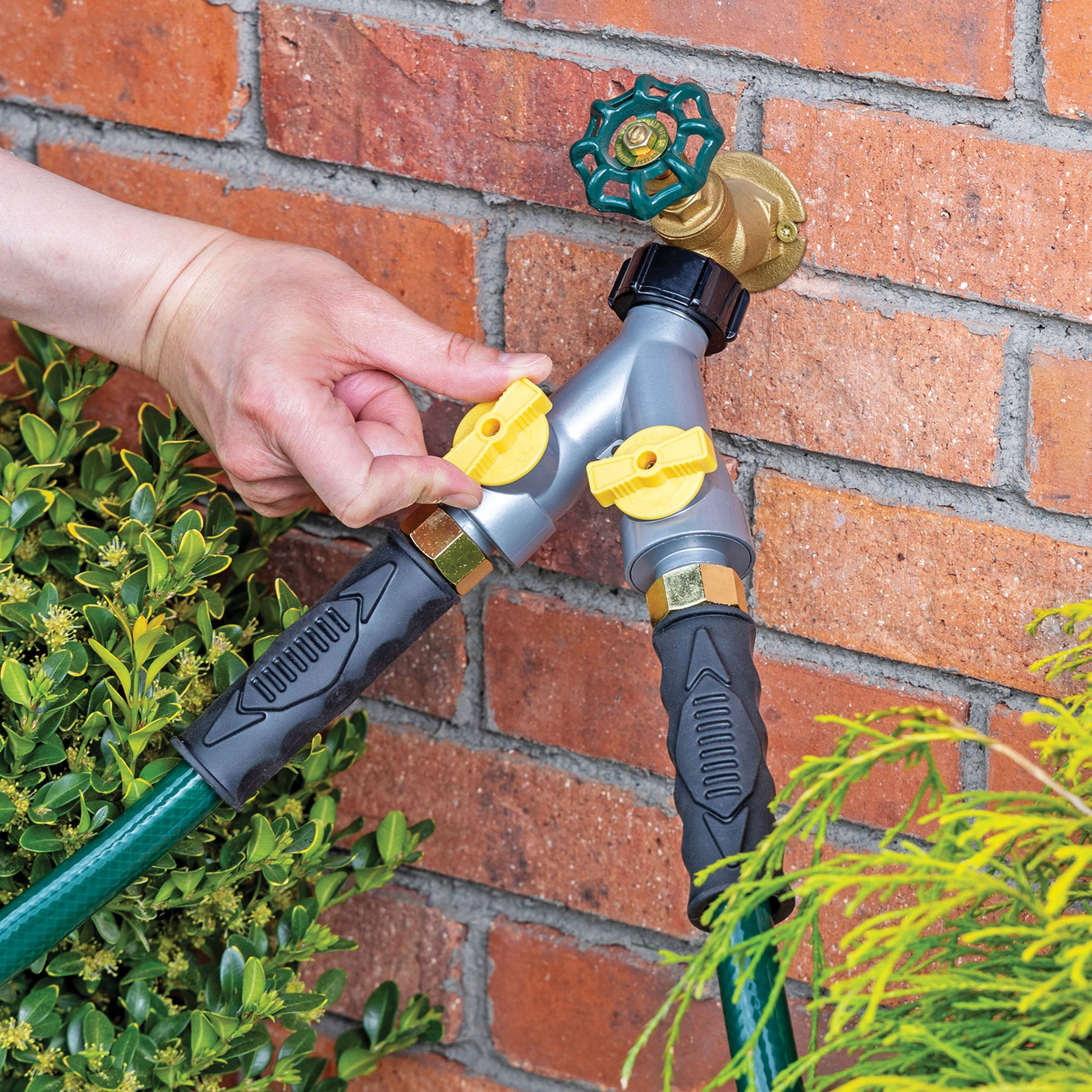 A Y split garden hose connector mounted on a red brick wall, featuring a gray body with a hand reaching out to turn one of two yellow valve controls .