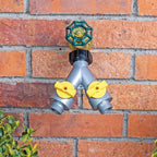 A Y split garden hose connector mounted on a red brick wall, featuring a gray body with two yellow valve controls and a green and gold main valve handle.