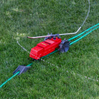 A red lawn sprinkler connected to a green hose is positioned on lush green grass, likely set to water the lawn with scattered water jets.