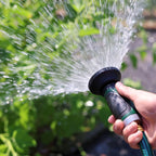 A person's hand holds a green and black garden hose with a nozzle, spraying a fine mist of water onto lush green plants in sunlight.