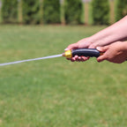 A person is holding a garden hose nozzle, spraying water onto grass, with a blurred green background indicative of a sunny garden or yard.