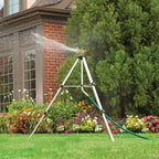 A tripod sprinkler is actively watering a lush garden with a variety of colorful flowers in front of a brick house with greenery.