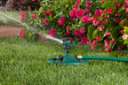 This image shows a green lawn sprinkler in action on a vibrant grassy area, with water spraying out amidst a backdrop of pink and red flowers.