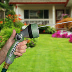 A person's hand is holding a garden hose with a spray nozzle, watering the lawn in front of a house with a porch and colorful flowerbeds.