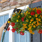 A person's hand is holding a hose with a spray nozzle, watering a vibrant hanging basket of red and yellow flowers in front of a house.