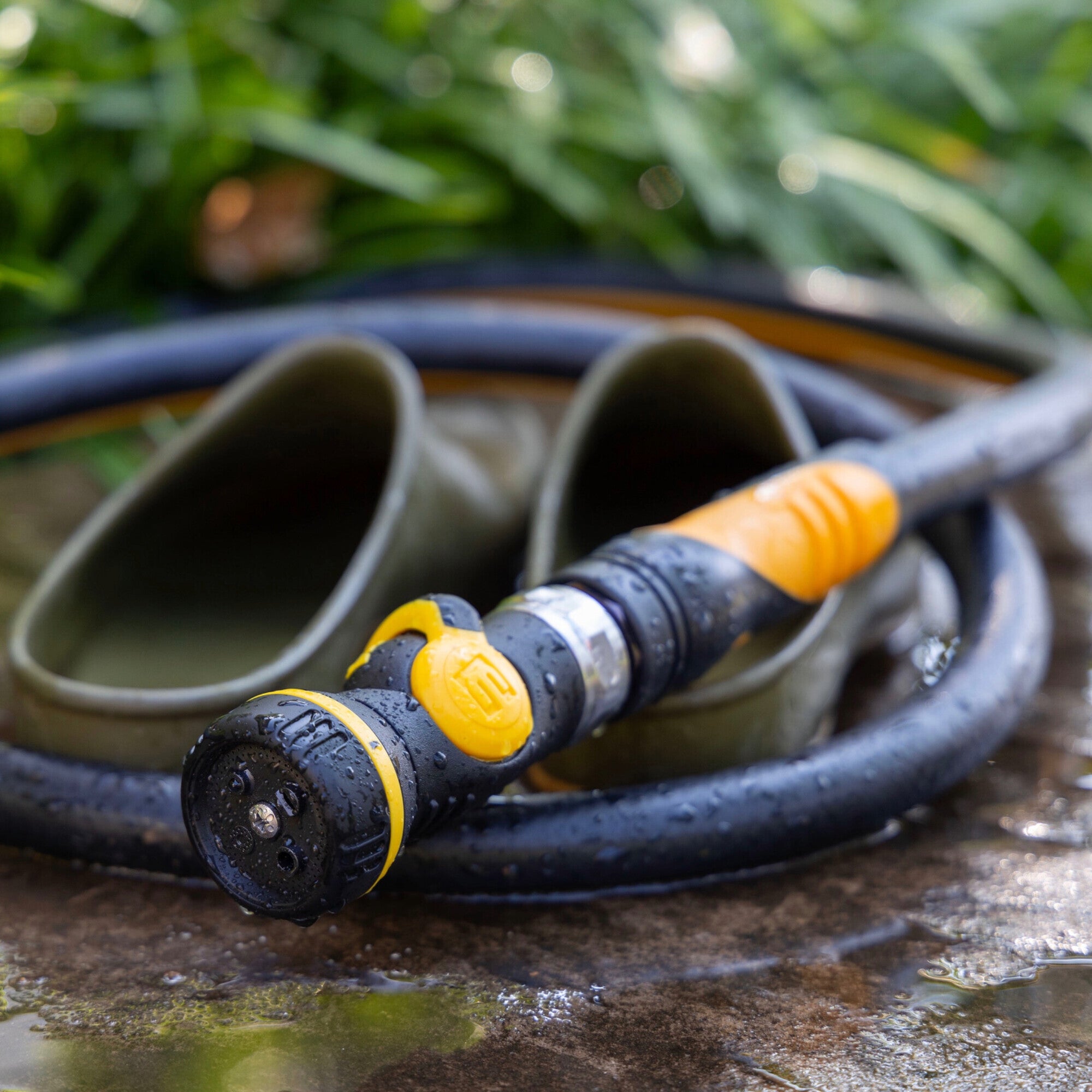 A close-up of an XT garden hose with the Elenor R901 Mini Nozzle resting on wet stone, with olive green rubber boots in the background and green grass above.