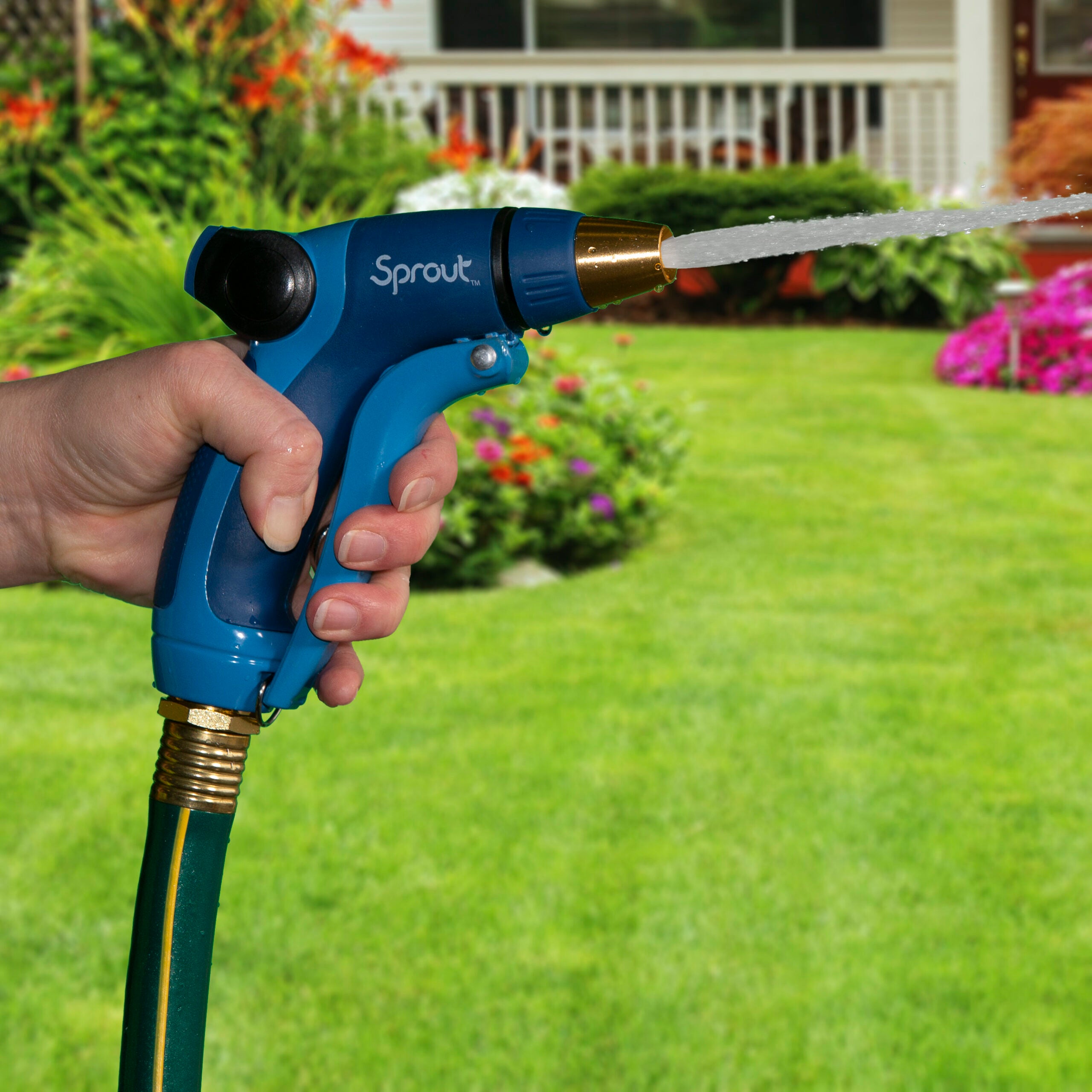 A person's hand holds a blue garden hose nozzle, spraying water onto a lush green lawn, with colorful flowers and a white fence in the background.