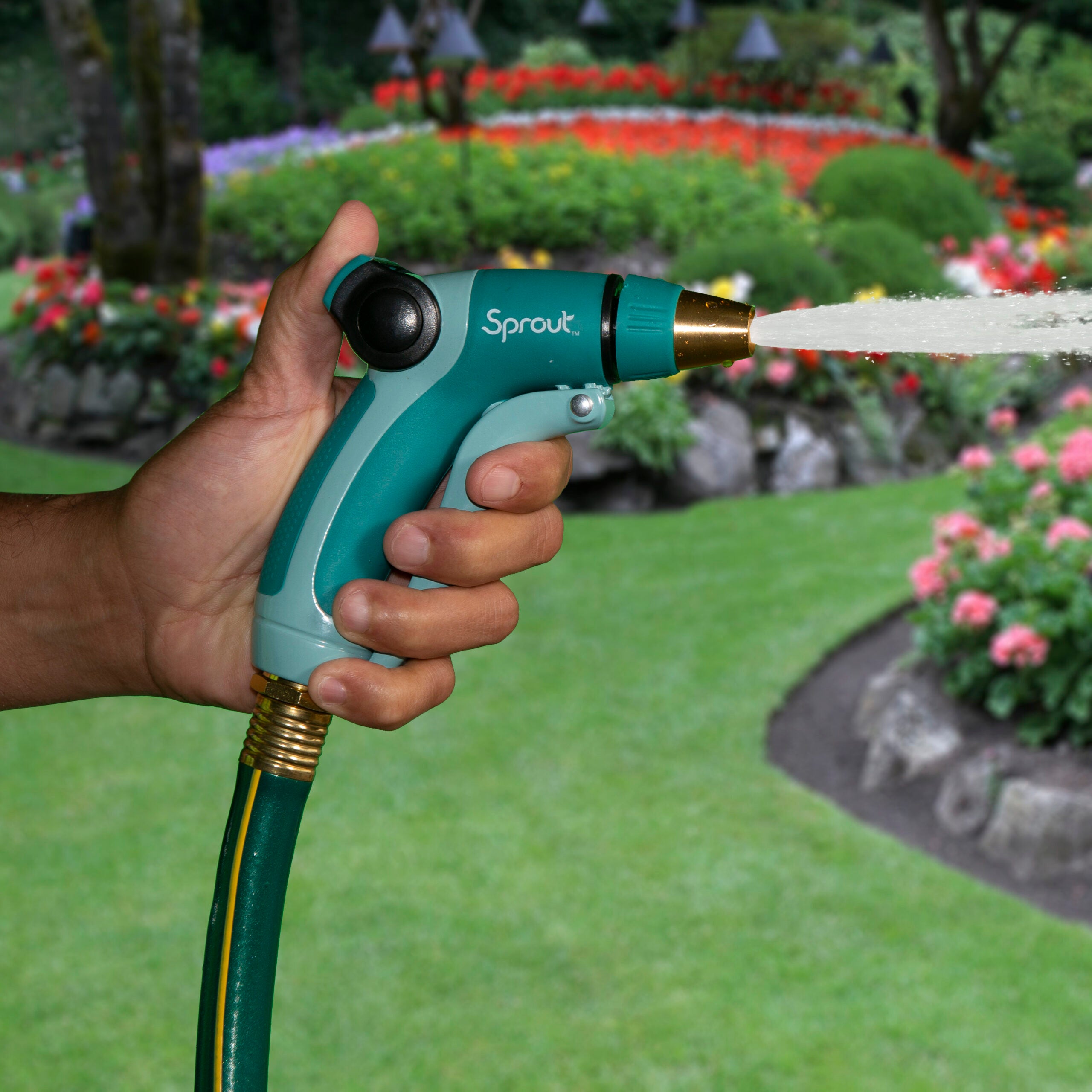 A person's hand is holding a green and black garden hose nozzle named "Sprout," spraying water, with a vibrant, colorful flowerbed and greenery in the background.