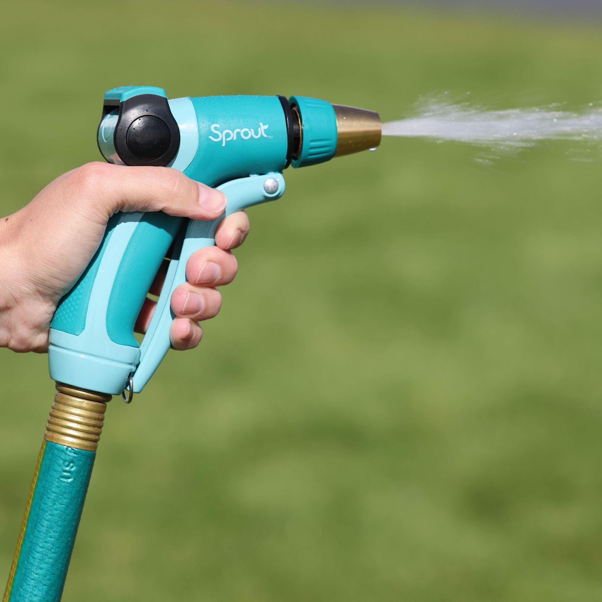 A person's hand is holding a blue and teal garden hose nozzle with the word "Sprout" on it, spraying water against a green grass background.