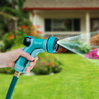 A person's hand is holding a blue and grey garden hose nozzle, spraying water onto a lawn with colorful flowers in front of a house.