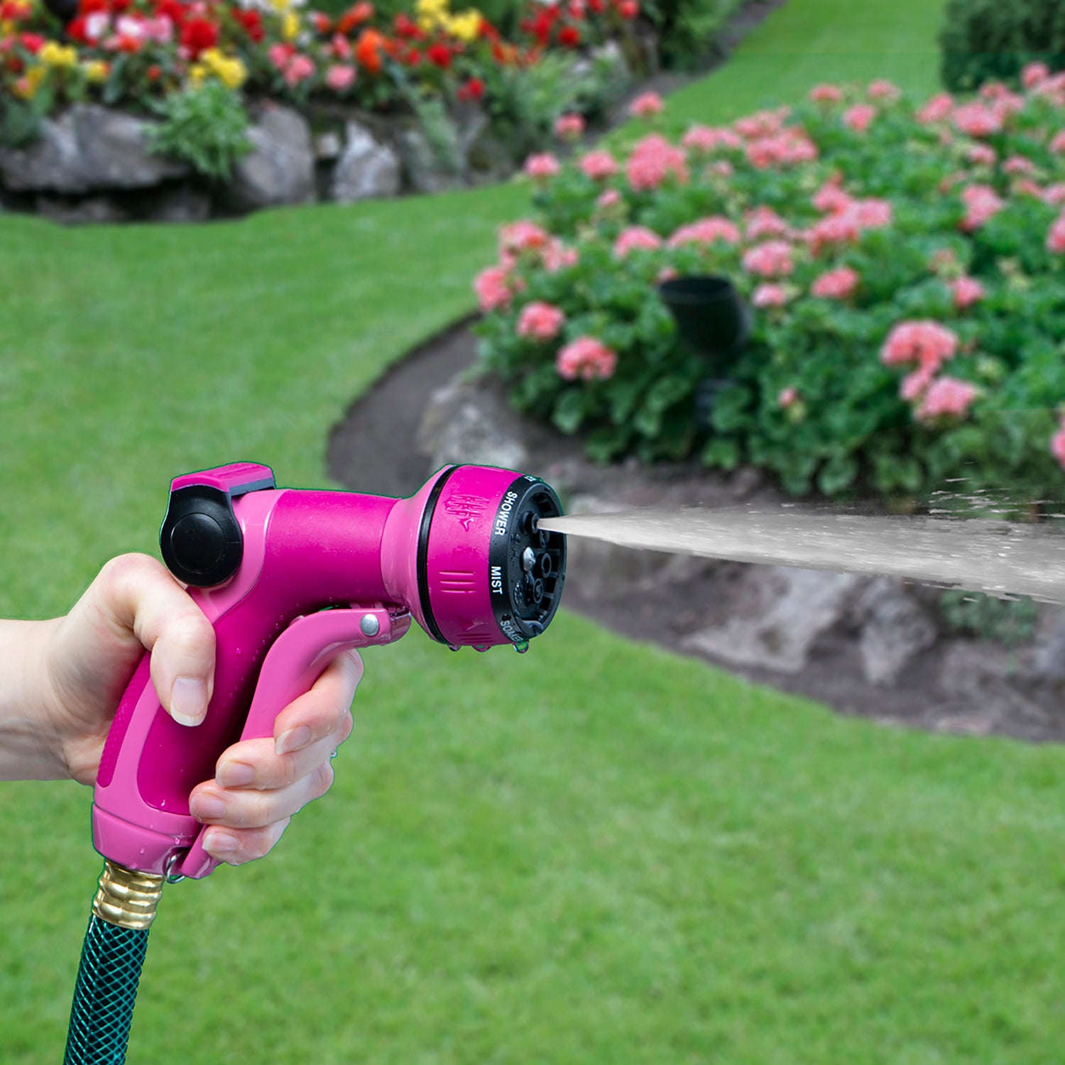 A person is holding a bright pink garden hose nozzle, spraying water onto a lush green lawn surrounded by vibrant pink and red flowers.