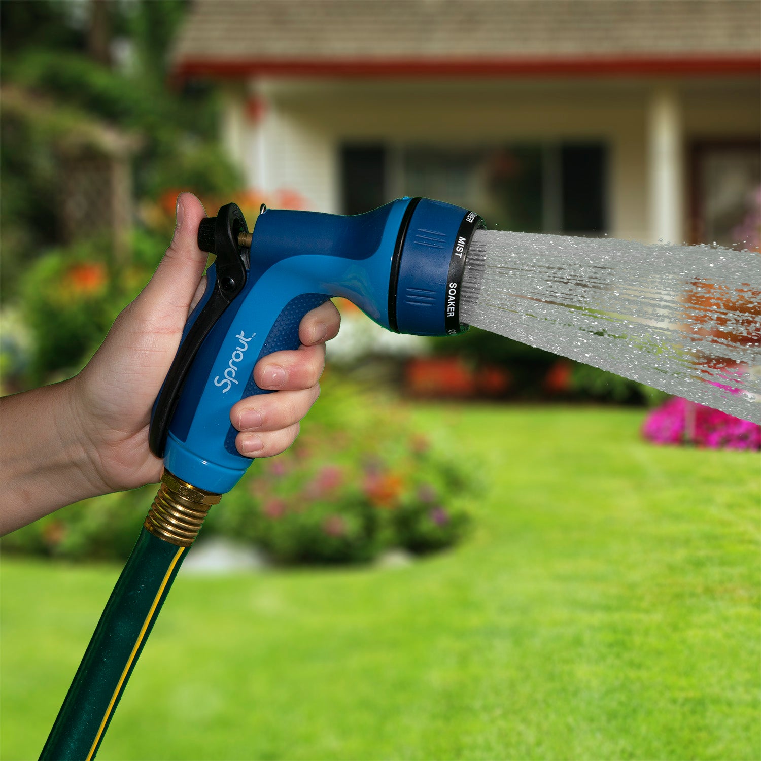A person's hand is holding a blue and black garden hose nozzle, spraying water onto a lawn. A residential house and garden are in the background.
