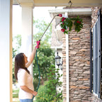 A person is watering a hanging flower basket with a hose in front of a house with stone accents. It's a sunny day with greenery around.
