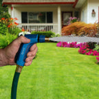 A person's hand is holding a blue hose nozzle, spraying water onto a lush green lawn with colorful flowerbeds in front of a white house.