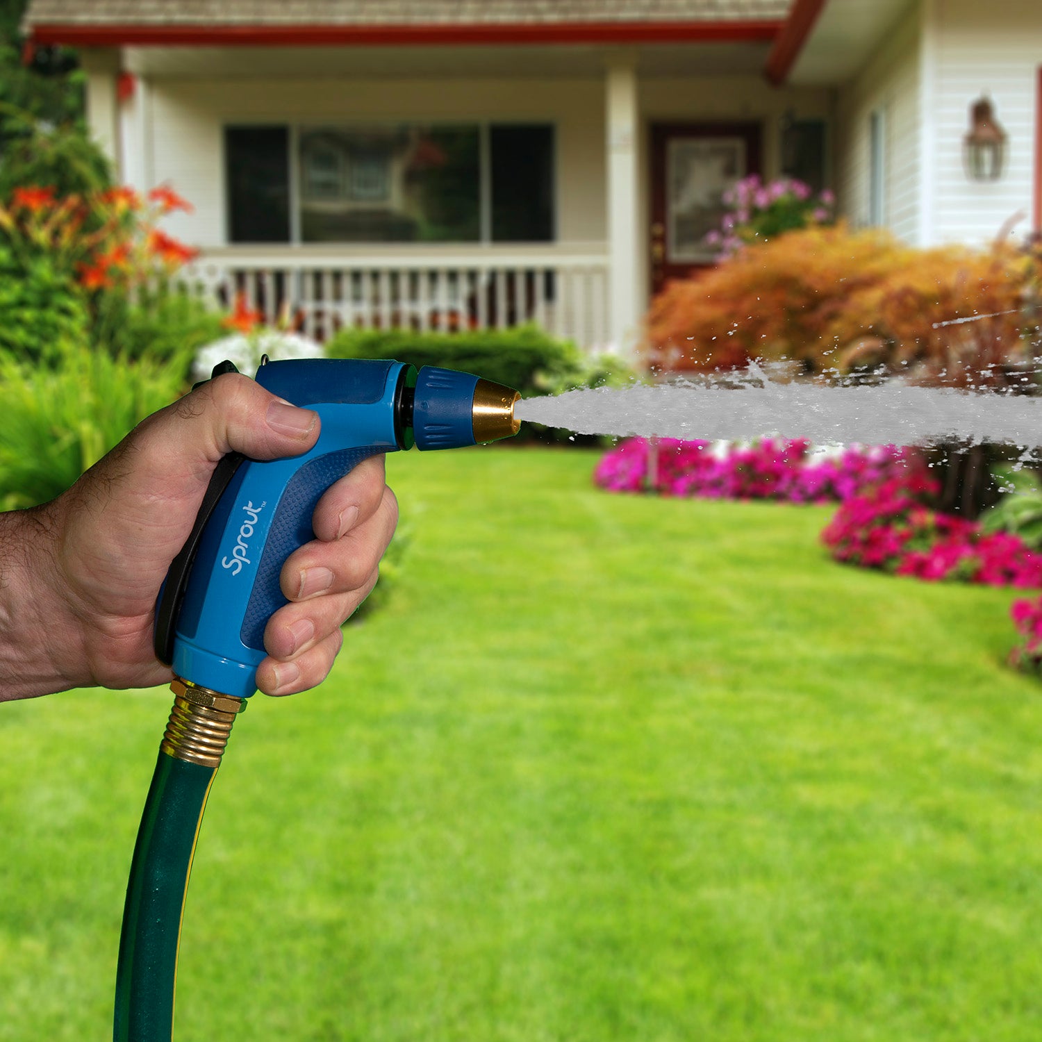 A person's hand is holding a blue hose nozzle, spraying water onto a lush green lawn with colorful flowerbeds in front of a white house.