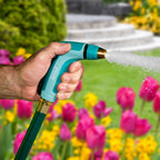 A person is holding a teal and black hose nozzle, spraying water in a vibrant garden with an abundance of pink tulips and a fountain.