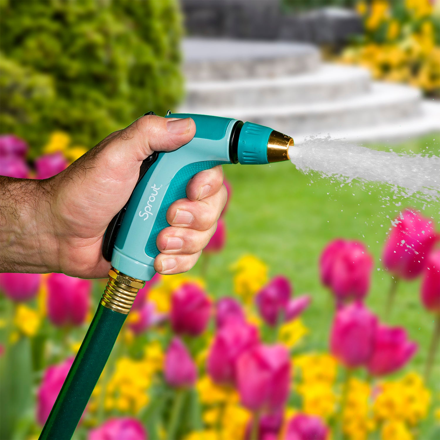 A person is holding a teal and black hose nozzle, spraying water in a vibrant garden with an abundance of pink tulips and a fountain.
