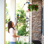 A person is watering hanging red flowers with a telescopic watering wand, standing on a porch by stone pillars and a black lantern.