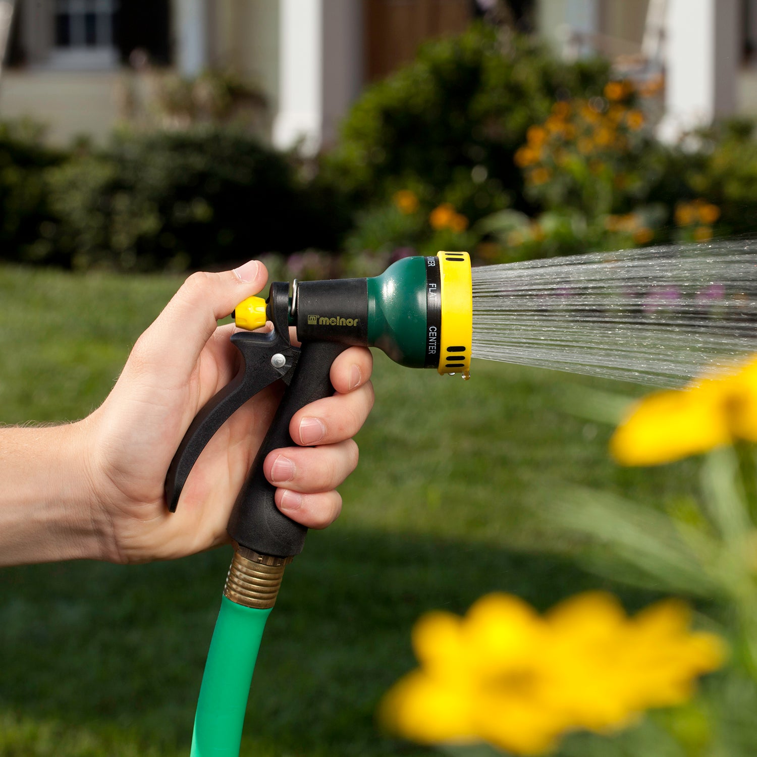 A person's hand is holding a green and yellow garden hose with a nozzle, spraying water, with a blurred background of a lawn and flowers.