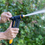 A hand using the Flow Control function on the Melnor XT300 Front-Trigger Adjustable Nozzle to strengthen water flow in front of a blurred garden background.