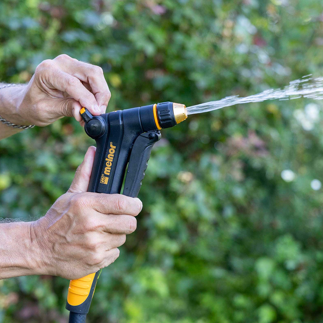A hand using the Flow Control function on the Melnor XT300 Front-Trigger Adjustable Nozzle to lessen water flow in front of a blurred garden background.