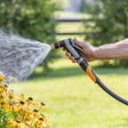 Hand holding the Melnor XT450 Heavyweight Metal Adjustable Nozzle spraying water on yellow flowers in a blurred garden background.