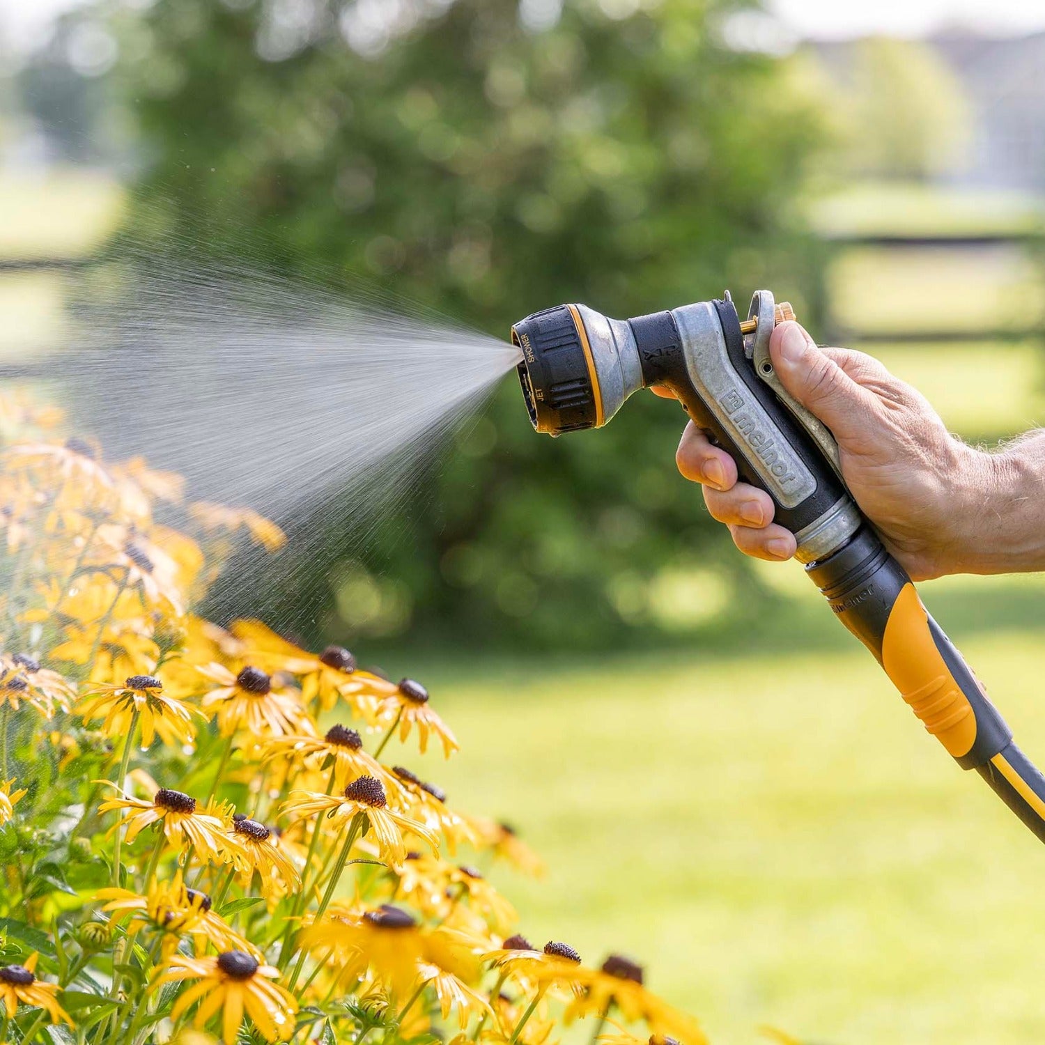 Hand holding the Melnor XT451 Heavyweight Metal 7-Pattern Nozzle spraying water on yellow flowers with a blurred green background.