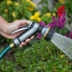 Hand holding the Melnor XT451 Heavyweight Metal 7-Pattern with water spraying, surrounded by greenery and flowers. 