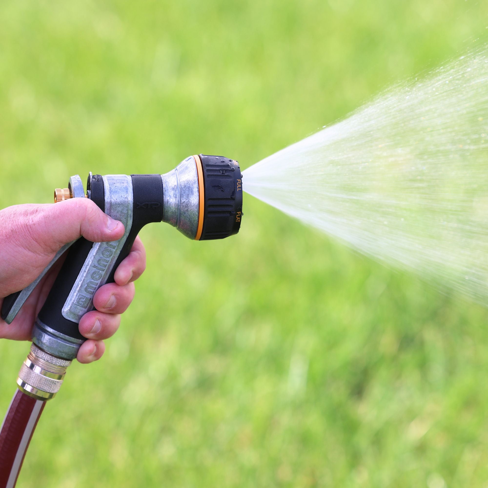Hand holding the Melnor XT451 Heavyweight Metal 7-Pattern Nozzle spraying water on a grassy background.