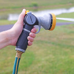 A person is holding a garden hose nozzle, ready to spray water. The nozzle is attached to a green and blue hose, set against a grassy background.