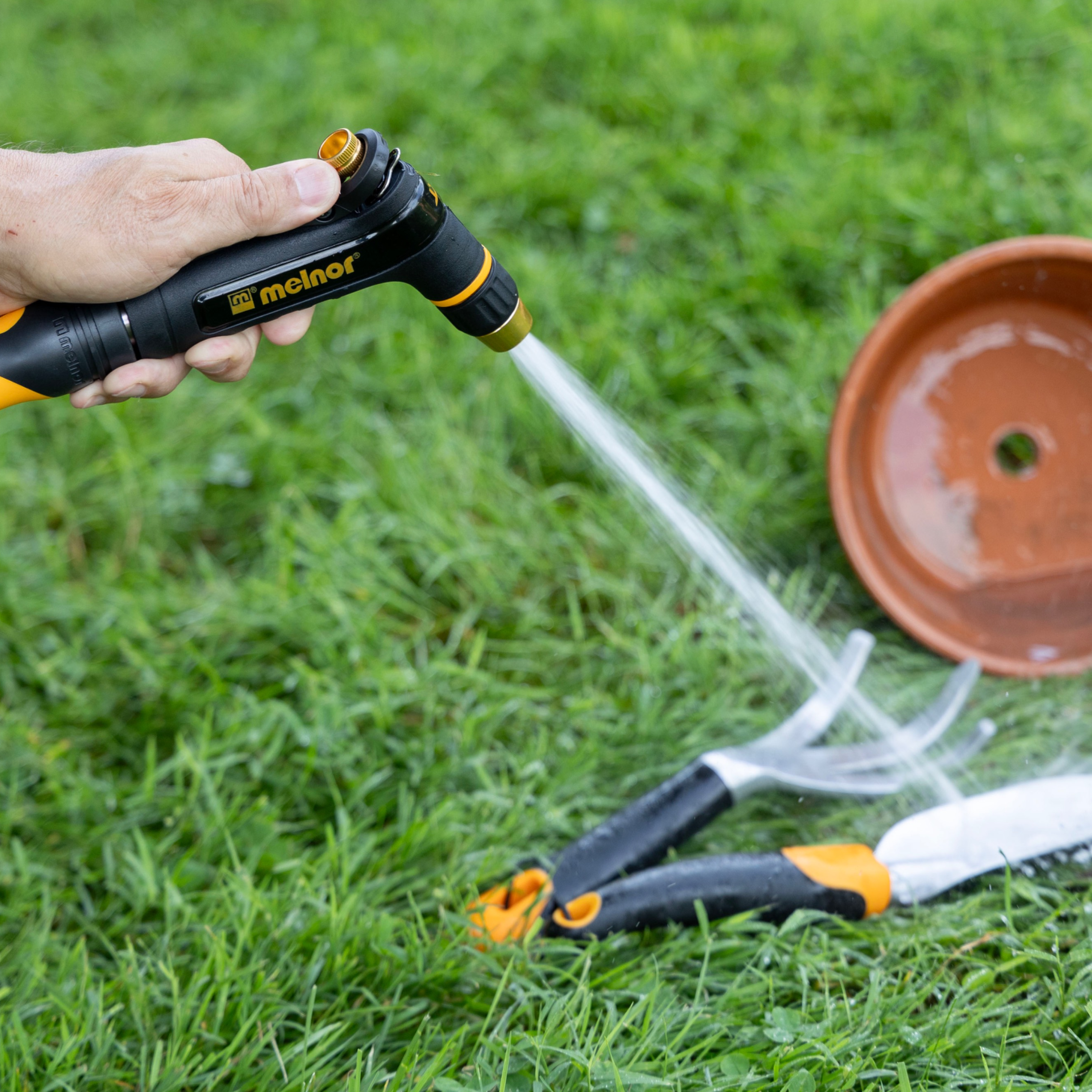 Melnor XT200 Rear Trigger Metal Adjustable Nozzle using the targeted jet spray setting on a gardening trowel and cultivator with a clay planting pot in the background on green grass.