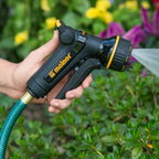 Hand holding the Melnor XT301 Front-Trigger Metal 8-Pattern Nozzle with plants in the background.