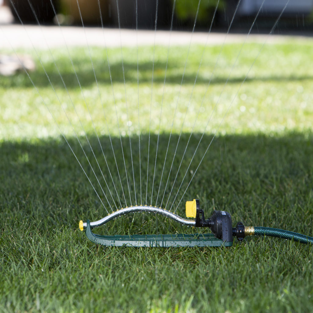 An oscillating lawn sprinkler is connected to a green garden hose, spraying water across lush, well-maintained grass under bright, sunny conditions.