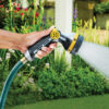A person's hand is holding a hose with a spray nozzle, watering plants in a vibrant garden. The nozzle has a yellow and black design.