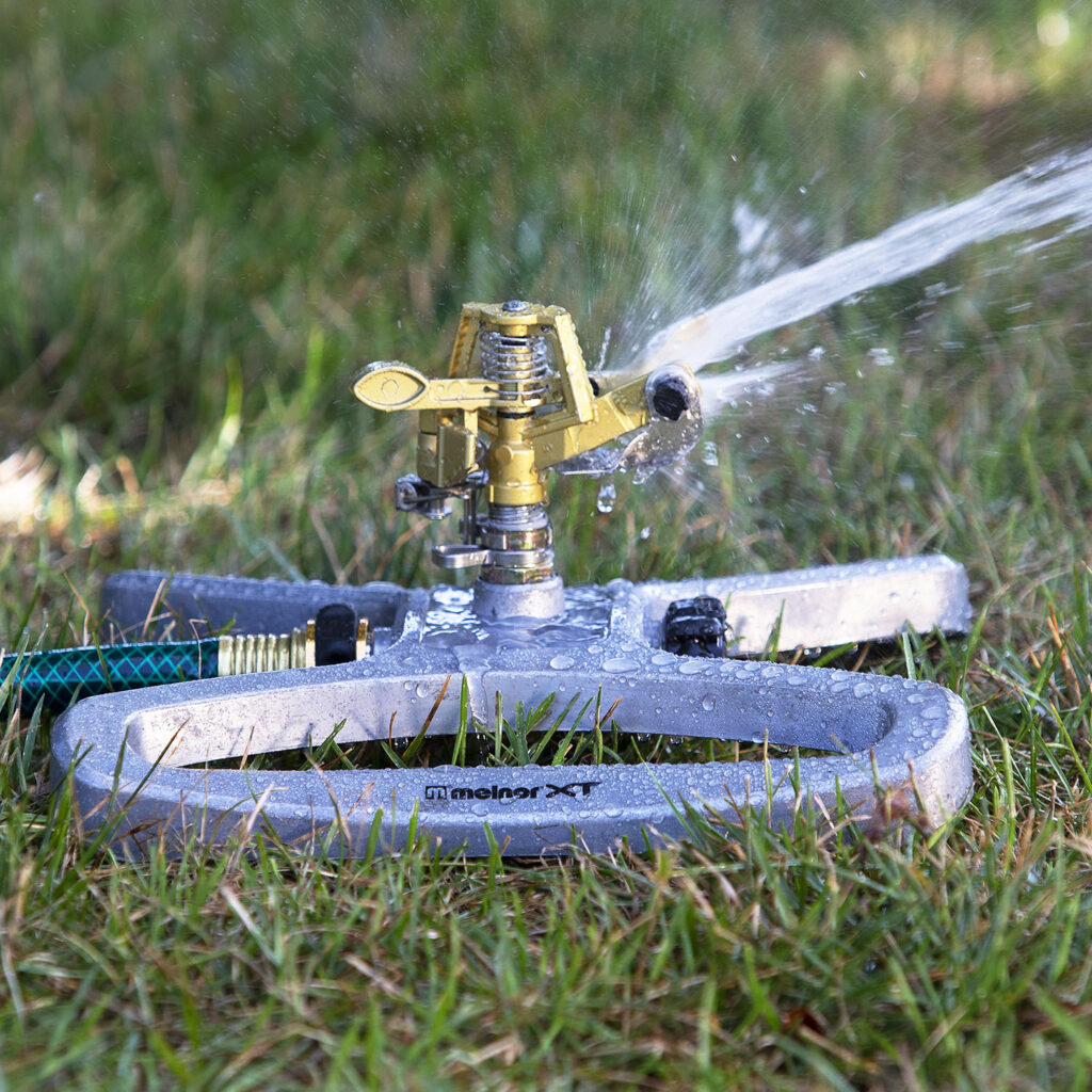 An oscillating sprinkler is actively watering the lawn, with jets of water spraying out in a fan shape. The grass appears well-maintained and green.